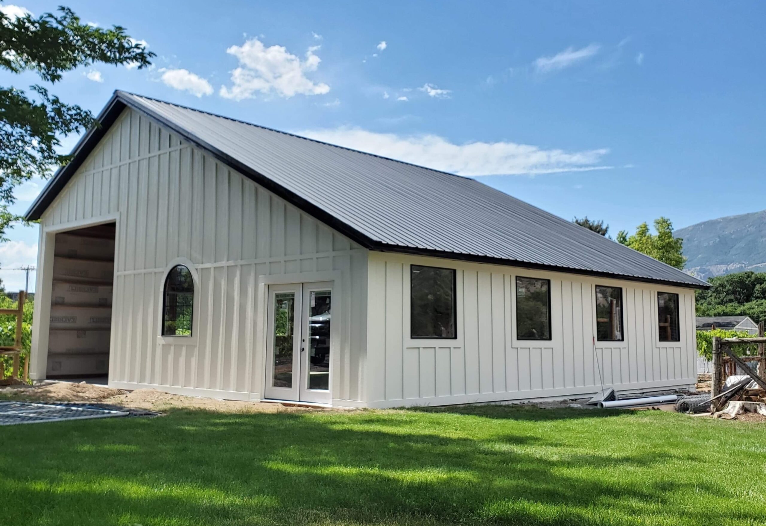 A white barn-style building with a black metal roof, large windows, and a double door, situated on a green lawn with a mountain view in the background.