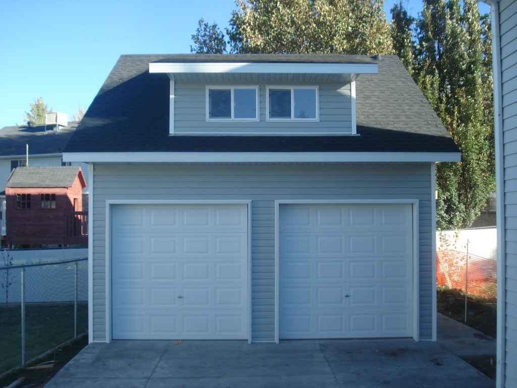 A two-car gray garage with white doors, featuring a dormer window on the black shingled roof, located in a residential backyard.