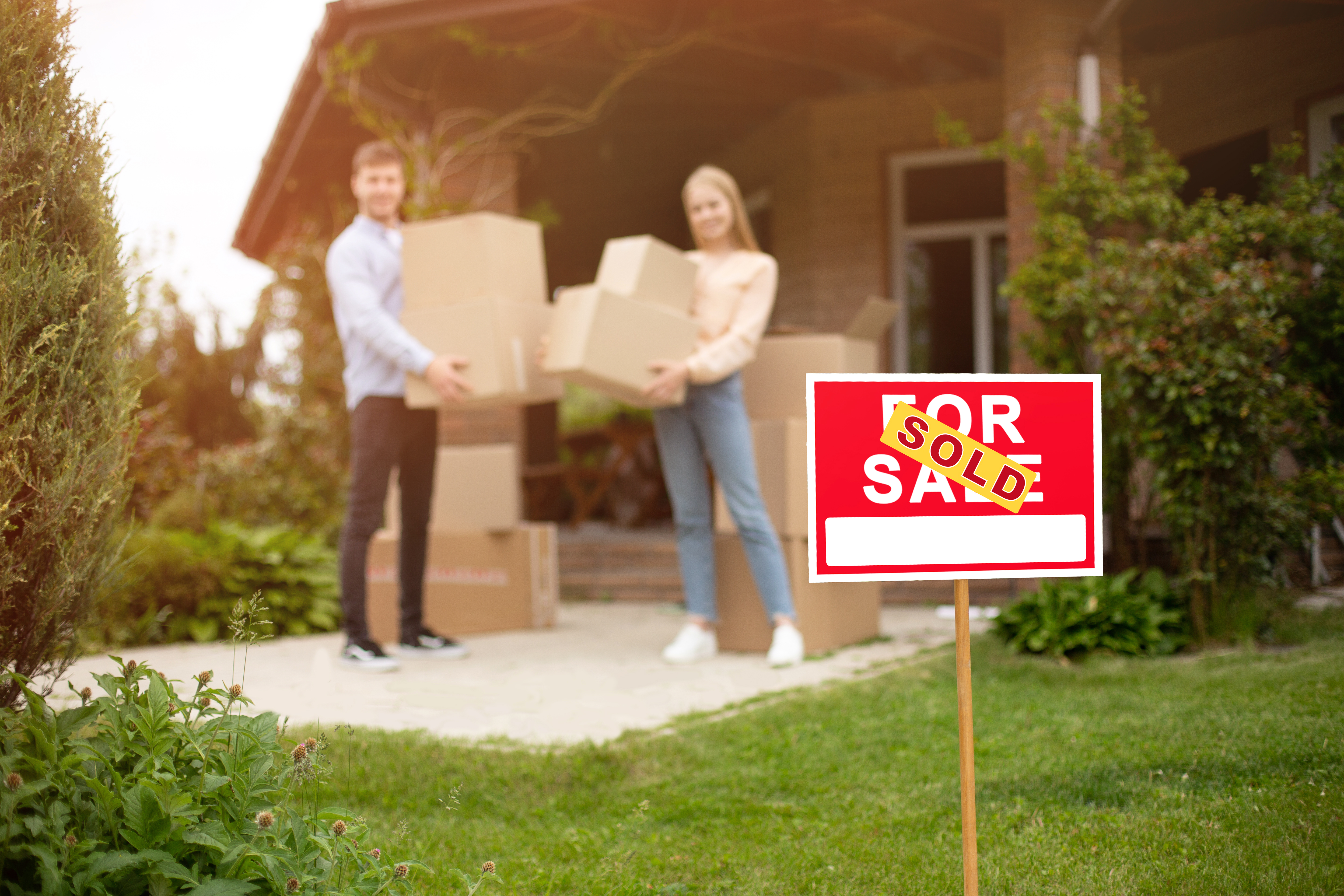 A "Sold" sign in front of a house with a young couple carrying moving boxes, symbolizing a recent home purchase.