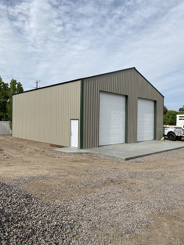 A large beige metal garage building with two white roll-up doors and a single side door, set on a concrete slab in a gravel area.