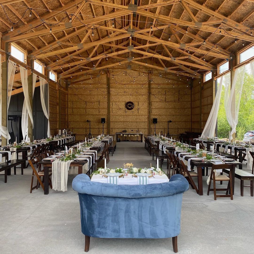 Interior of a pole barn set up for an event, with long dining tables, candles, greenery, string lights, and a blue couch at the front.