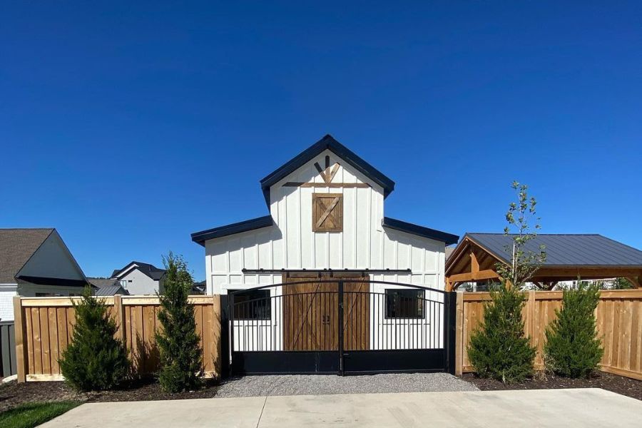 Small white barn-style building with a black gate, wooden accents, and a fenced yard, set against a clear blue sky.