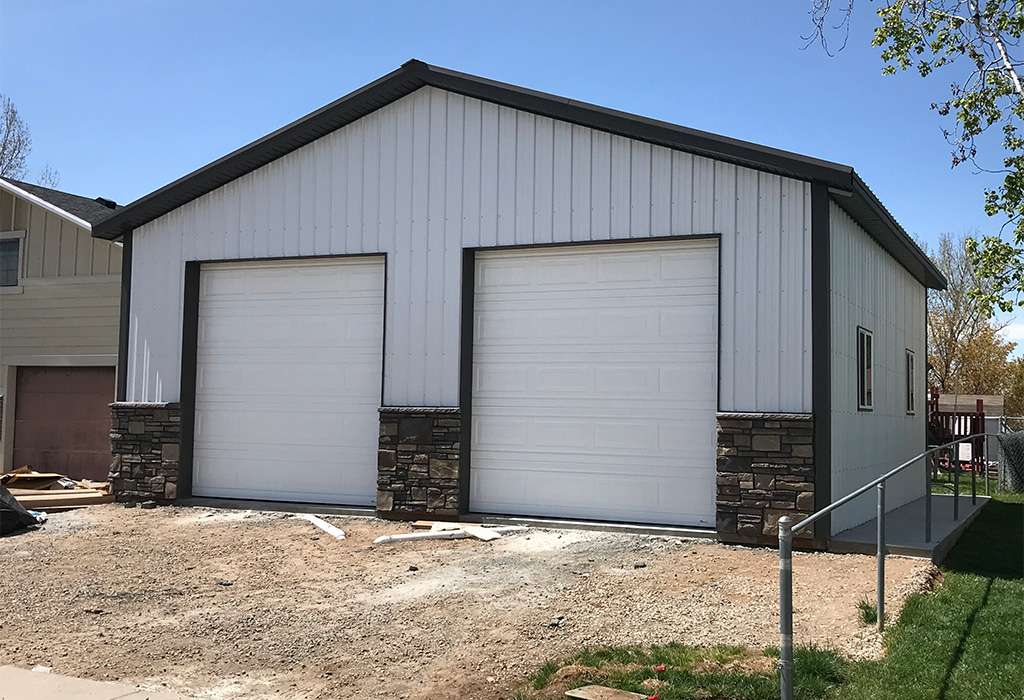 White pole barn building with two large garage doors and stone accents along the bottom, set in a residential area.