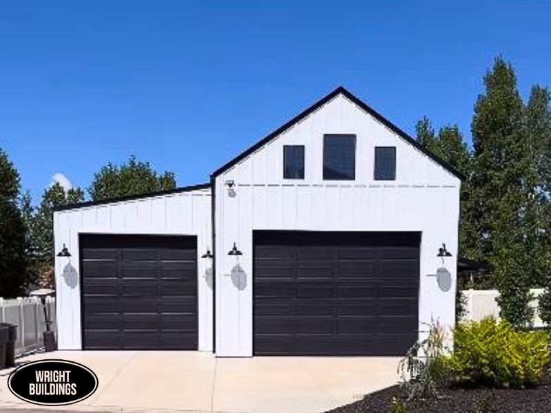 A modern white pole barn-style building with two black garage doors, a gable roof, and three clerestory windows, surrounded by greenery and a paved driveway.