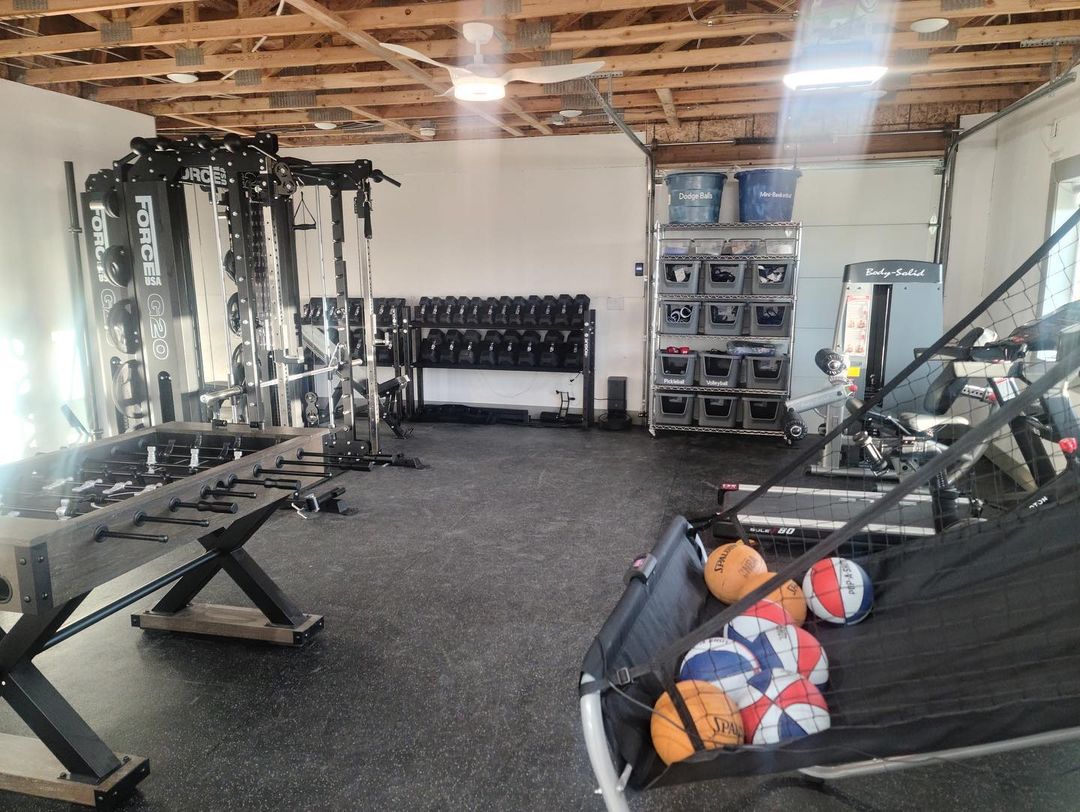 A home gym inside a pole barn featuring weight racks, a multi-functional fitness machine, storage bins, and sports equipment, with an exposed wood ceiling.