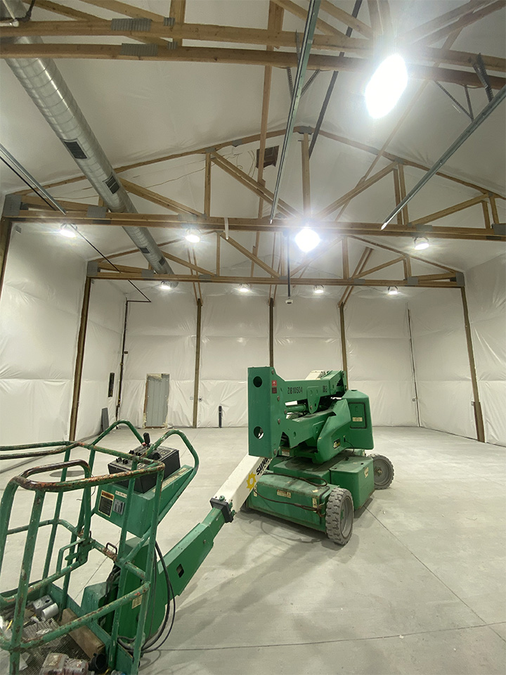 Interior of a steel building under construction, featuring exposed wooden trusses, insulated walls, and green construction equipment on a concrete floor.