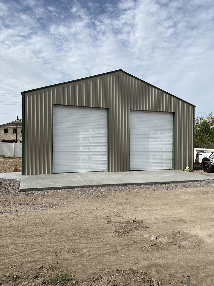 A tan metal shop building with two large white garage doors, set on a concrete foundation under a partly cloudy sky.