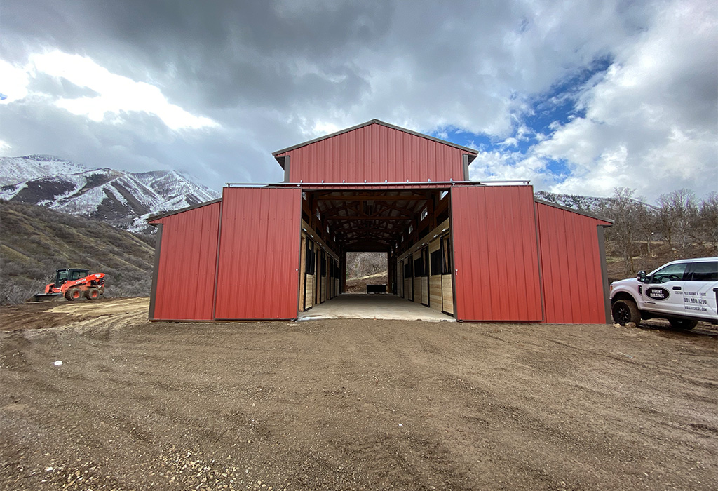 A red horse barn with a central aisle barns, surrounded by mountainous terrain and a cloudy sky, showcasing sturdy construction and a spacious layout.