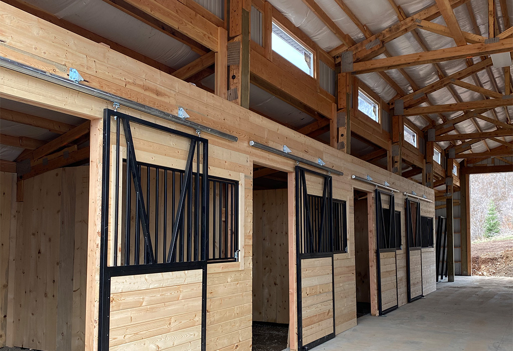 Interior view of a horse barn featuring wooden stalls with metal sliding doors and ample natural light from upper windows.