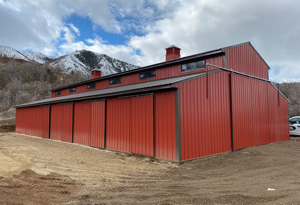 Red metal horse barn with a durable exterior, featuring ventilation cupolas and large sliding doors, set against a mountain backdrop.