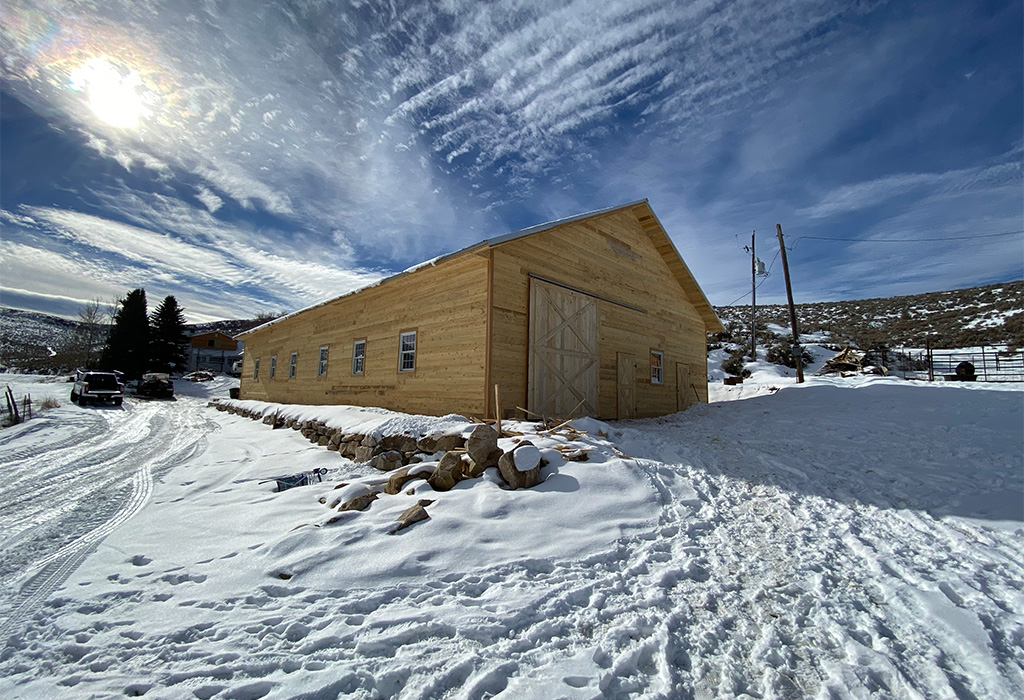 Wooden post frame barn in a snowy rural landscape with a clear blue sky.