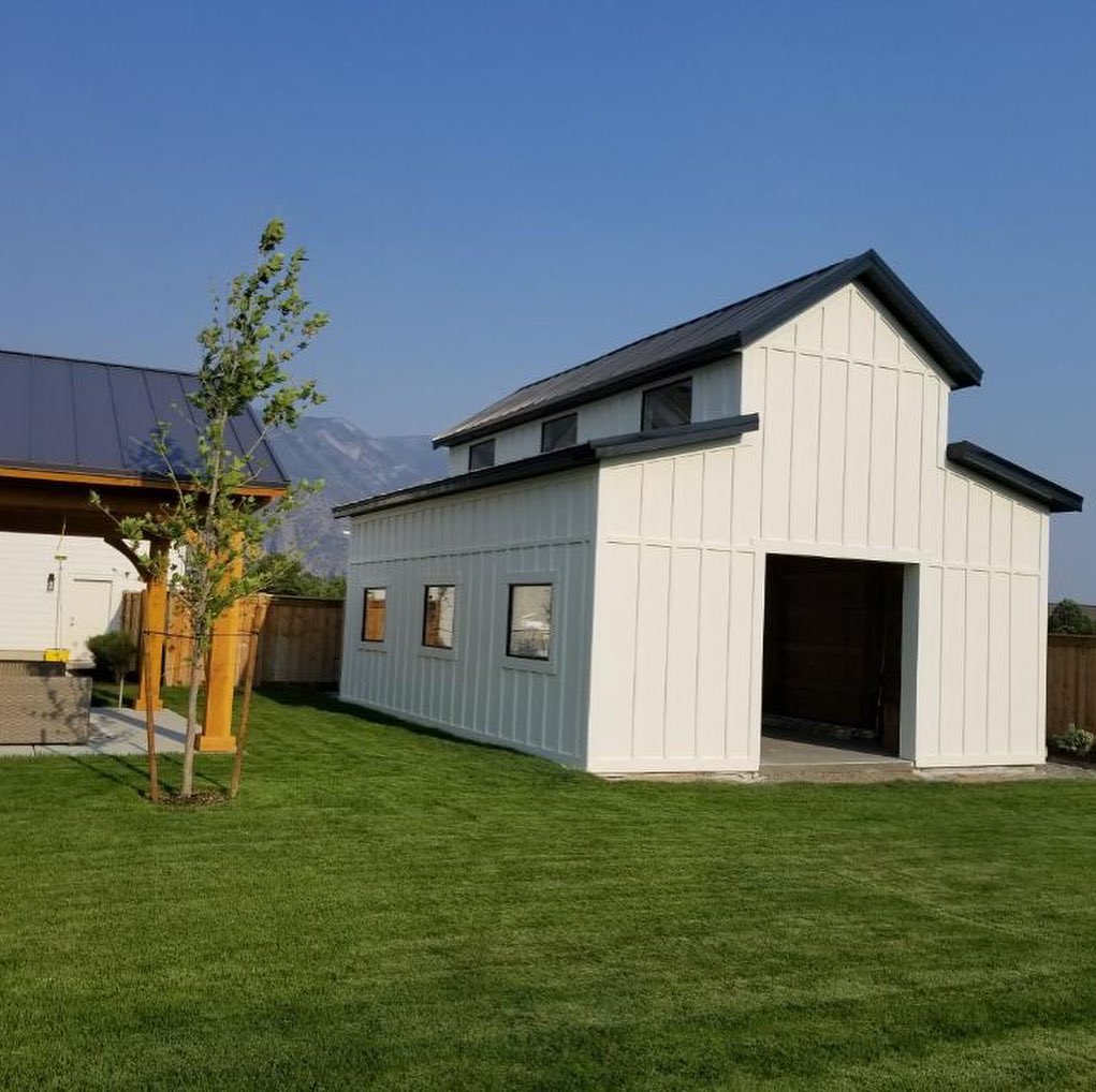 A white barn-style structure with black roofing, surrounded by a lush green lawn and set against a clear blue sky and mountain backdrop.