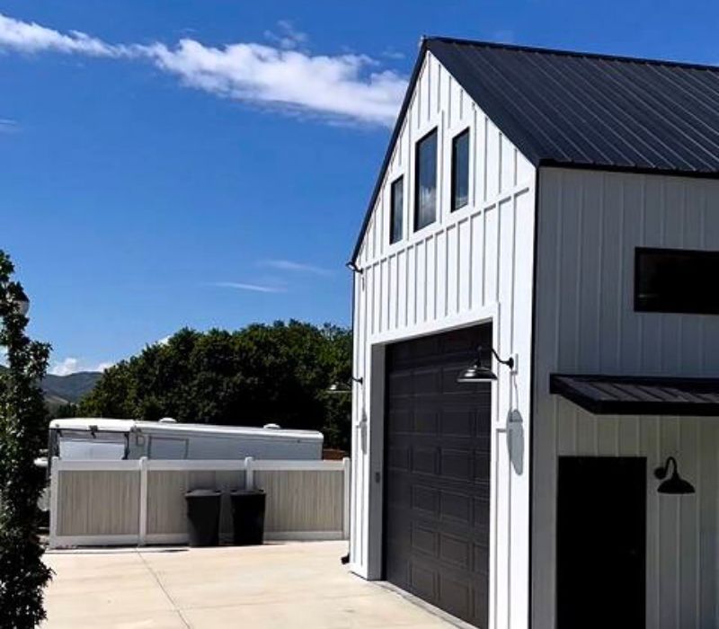 A modern white barn-style garage with black accents, large windows, and a spacious driveway, set against a backdrop of greenery and blue skies.