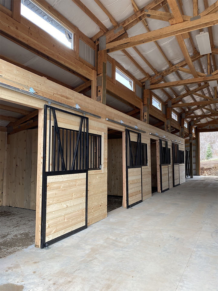 Interior view of a wooden horse barn with spacious stalls, sliding doors, and natural light from windows above.