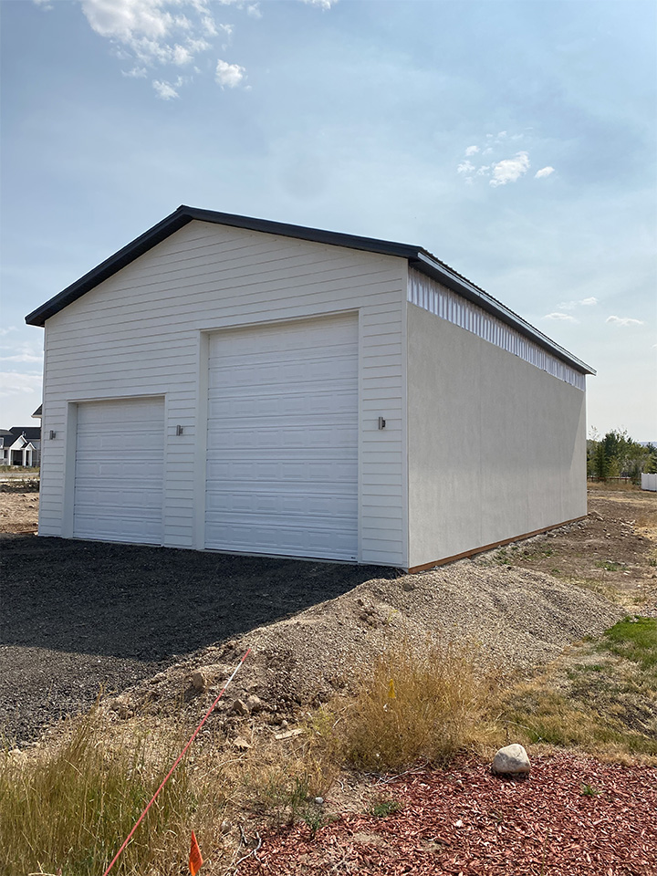 Large white detached garage with two roll-up doors, set on a gravel lot under a bright sky.