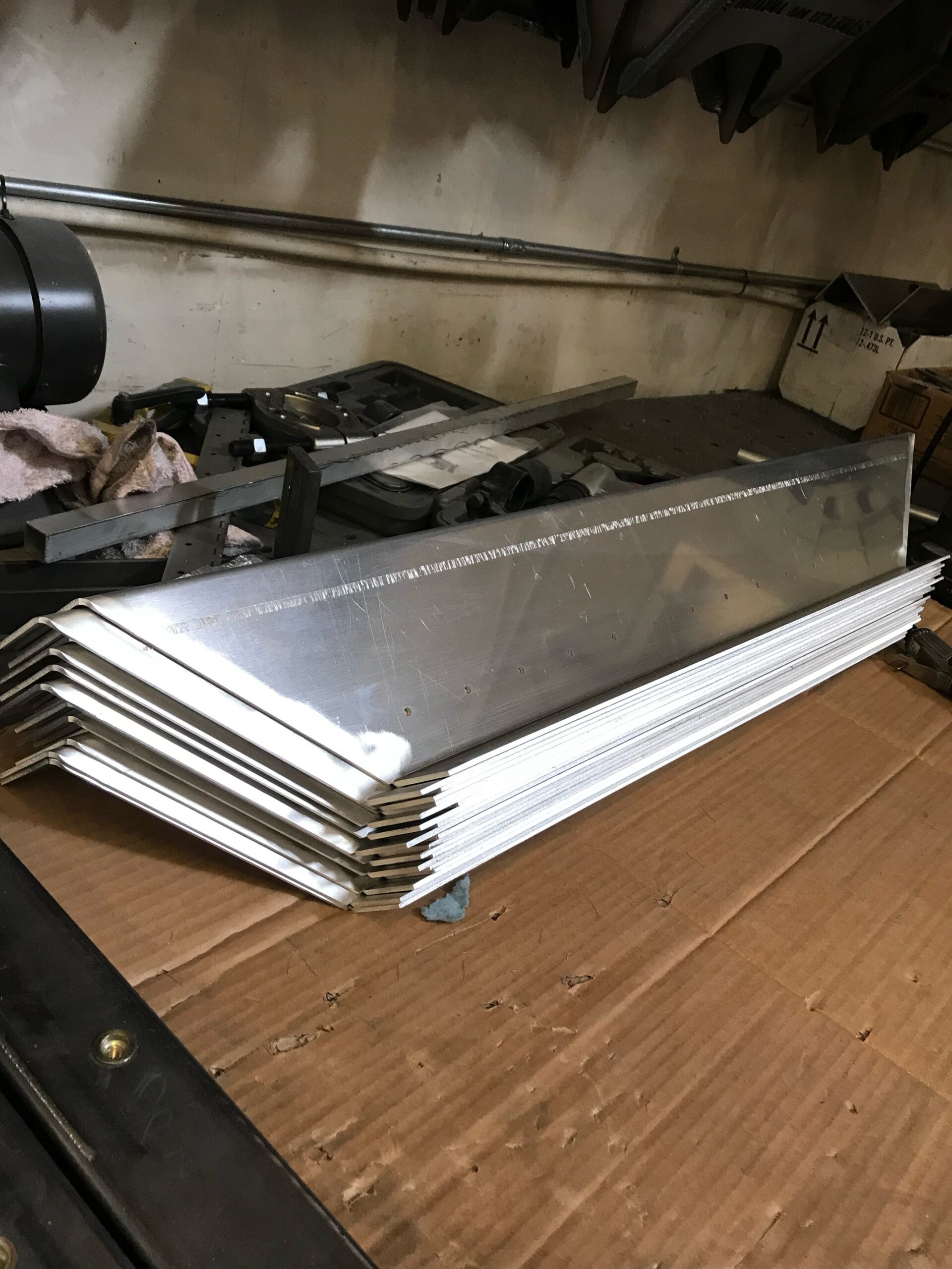 A stack of bent sheet metal pieces on a cardboard surface in a workshop with tools and materials nearby.