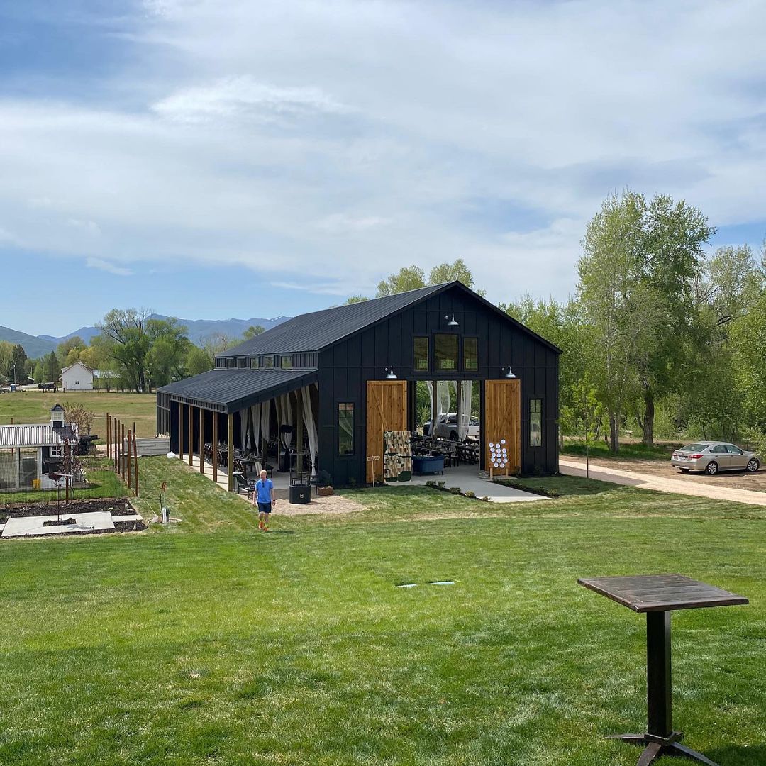 A sleek black pole barn with wooden sliding doors, open sides, and a lush green lawn in a rural setting.